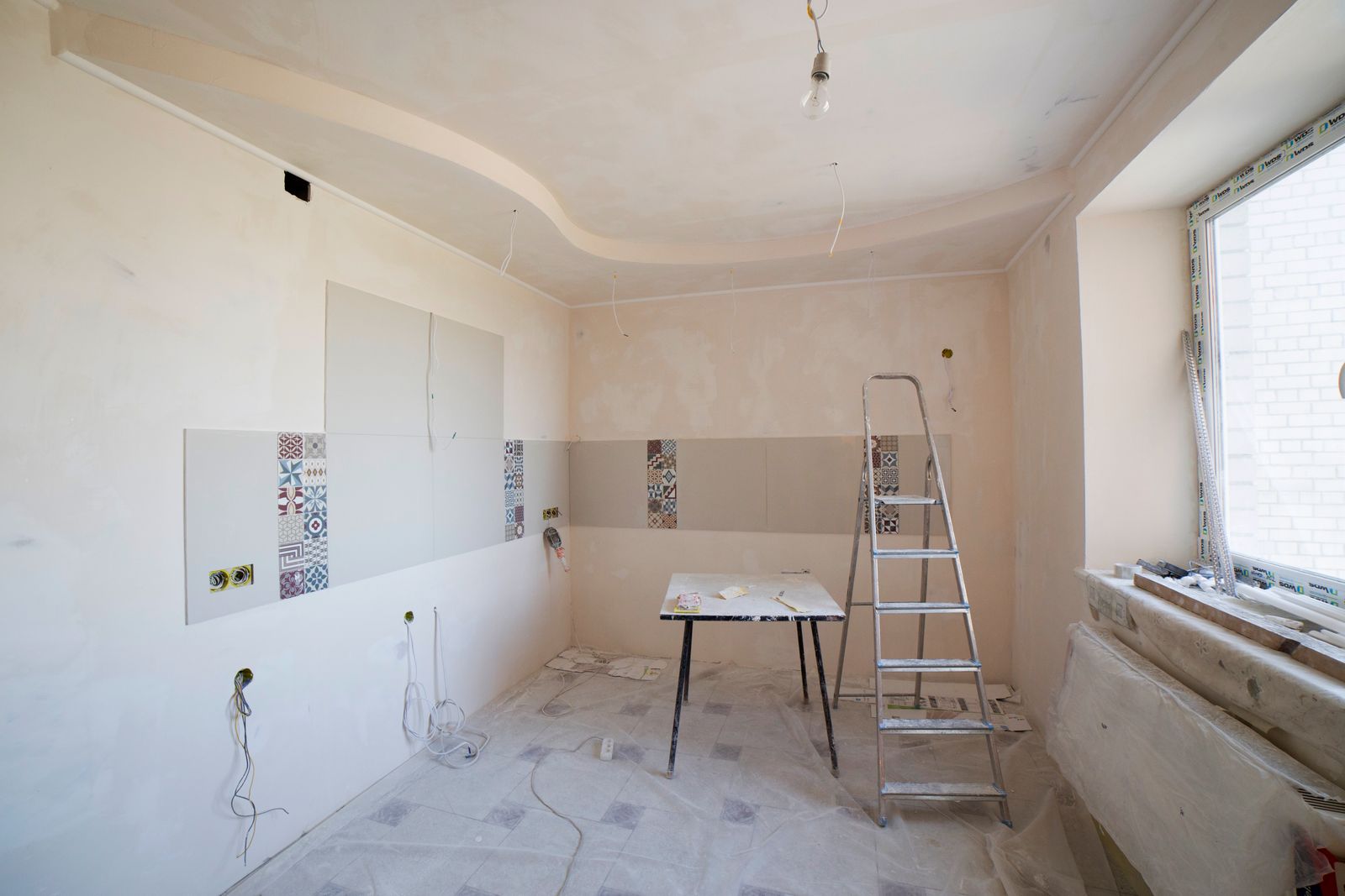 Cocina en obras: paredes a medio alicatar, escalera, mesa y cables sueltos.